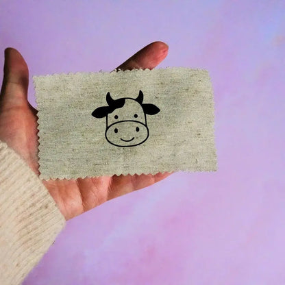 Main tenant une étiquette en tissu beige avec un motif de tête de vache noir minimaliste, sur fond lavande.