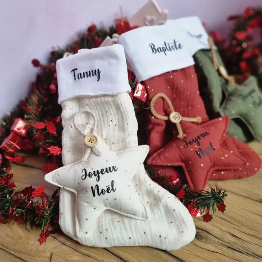 Etoiles et bottes de Noël à suspendre à tissu double gaze de coton blanc (au premier plan), rouge (au second plan) et vert (à l'arrière plan). Les bottes de Noël sont personnalisées avec un prénom floqué en noir sur leur partie supérieure en tissu polaire blanc. Les étoiles de Noël, quant à elles, sont personnalisées avec l'inscription "Joyeux Noël".