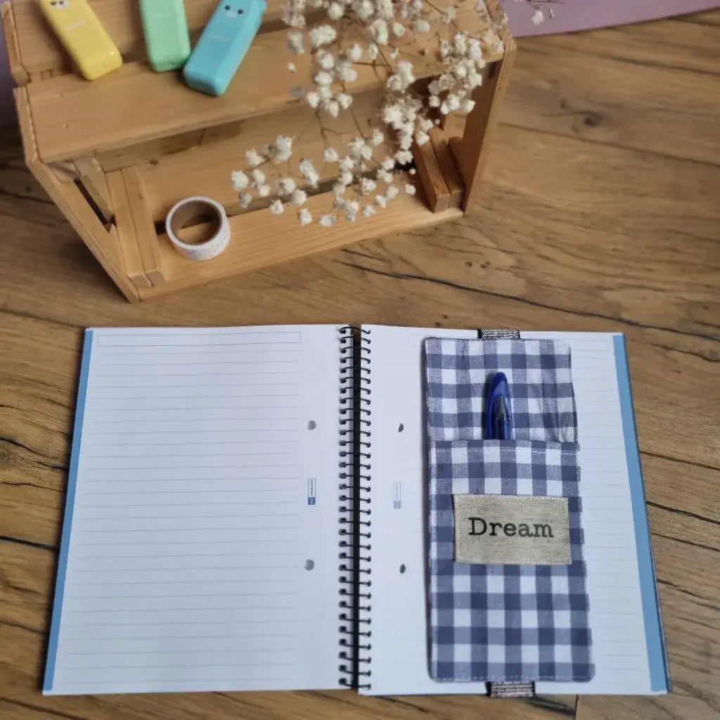 Étui à stylos en tissu vichy bleu à fixer sur la couverture d’un carnet A5. Pratique et nomade, il permet de garder vos stylos toujours à portée de main. Inscription "Dream" floquée sur une étiquette en tissu.