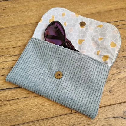 Etui à lunettes en velours côtelé de coloris bleu clair. Cet étui est ouvert. Une paire de lunettes de soleil est à moitié glissée à l'intérieur. On distingue la doublure de cet étui à lunettes en coton imprimé à motif argéo.