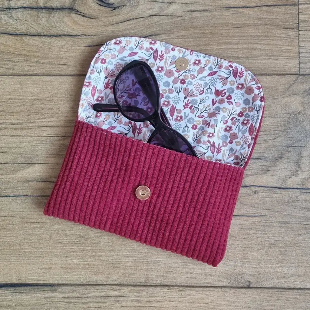 Étui à lunettes en velours côtelé bordeaux avec doublure fleurie et bouton pression, posé sur du bois.