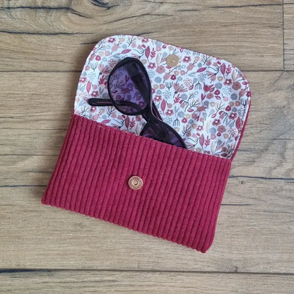 Étui à lunettes en velours côtelé bordeaux avec doublure fleurie et bouton pression, posé sur du bois.