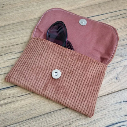 Etui à lunettes en velours côtelé de coloris marron. Cet étui est ouvert. Une paire de lunettes de soleil est à moitié glissée à l'intérieur. On distingue la doublure de cet étui à lunettes en coton imprimé à motif uni terracotta.