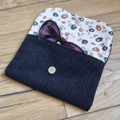 Etui à lunettes en velours côtelé de coloris noir. Cet étui est ouvert. Une paire de lunettes de soleil est à moitié glissée à l'intérieur. On distingue la doublure de cet étui à lunettes en coton imprimé à motif léopard.