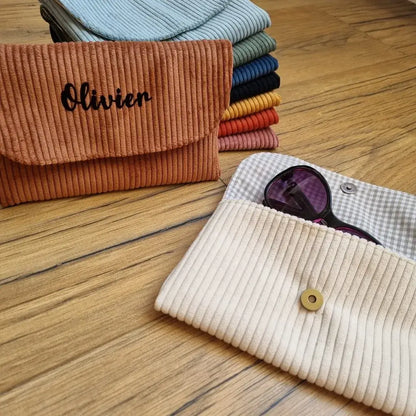 Au premier plan, un étui à lunettes en velours côtelé beige est posé au sol. Cet étui est ouvert. Une paire de lunettes de soleil est à moitié glissée à l'intérieur. On peut distinguer la doublure de cet étui en coton imprimé à motif carreaux beiges. A l'arrière plan son empilés des étuis du même type, également en velours côtelé de coloris divers. Devant cette pile est posé un étui à lunettes en velours côtelé marron personnalisé avec le prénom "Olivier" floqué en noir sur son rabat.