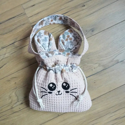 Pochon rose en nid d'abeille avec flocage chat noir et oreilles fleuries, posé sur un parquet en bois clair.