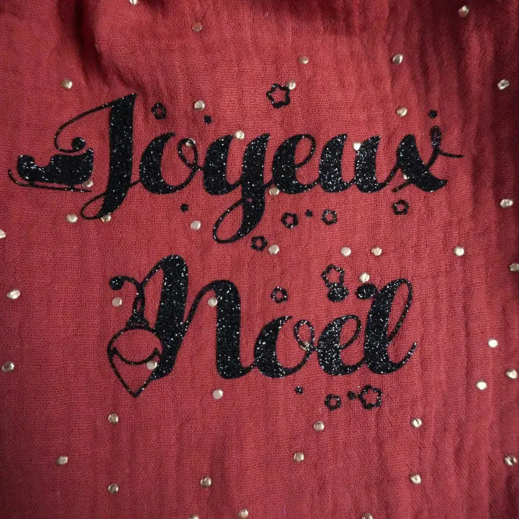 Inscription "Joyeux Noël" en flocage noir pailleté sur tissu rouge en double gaze, décorée de petits pois dorés scintillants et de motifs étoiles. Cette création festive artisanale évoque la magie de Noël et la chaleur des fêtes. Idéale pour illustrer un pochon, une décoration textile ou un emballage cadeau personnalisé fabriqué à la main pour les fêtes de fin d’année.