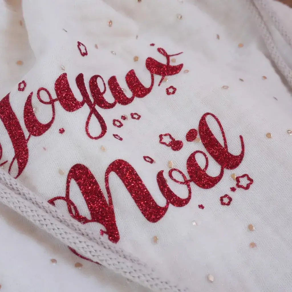 Flocage pailleté rouge “Joyeux Noël” appliqué sur tissu blanc en double gaze, orné de petits pois dorés et d’étoiles scintillantes. Ce visuel met en avant un motif festif et raffiné, parfait pour un pochon ou une décoration textile artisanale. Idéal pour illustrer une création de couture personnalisée aux accents de Noël, combinant brillance, douceur et esprit fait main pour les fêtes de fin d’année.