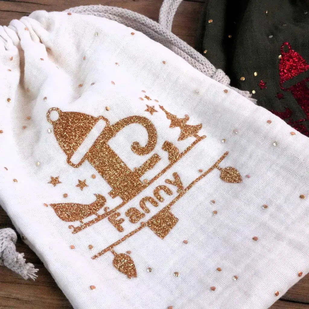 Flocage personnalisé en flex thermocollant doré pailleté représentant une initiale F stylisée et le prénom Fanny sur une pochette en double gaze blanche. Décor scintillant idéal pour customiser un pochon, une trousse ou un sac de Noël fait main. Effet brillant durable pour une création textile élégante et festive.