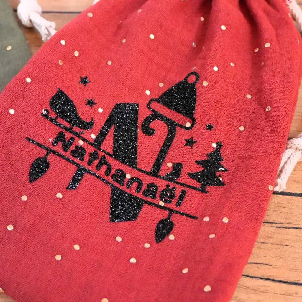 Pochon rouge en double gaze décoré d’un flocage personnalisé noir pailleté avec initiale et prénom Nathanaël. Motif festif composé d’un bonnet de Noël, d’un sapin et d’une guirlande lumineuse, réalisé en flex thermocollant brillant. Idéal pour personnaliser un sac cadeau ou une pochette de fête faite main.