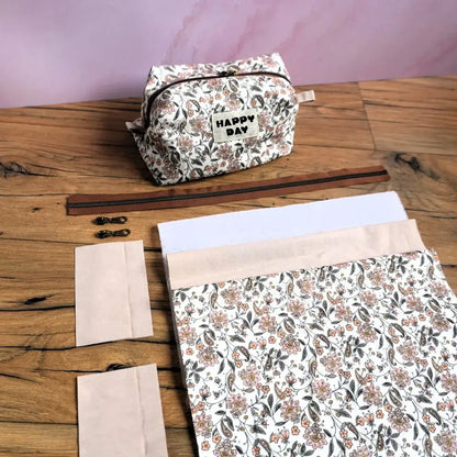 Kit couture complet pour réaliser une trousse carrée à motif fleuri, comprenant tissu imprimé, zip marron, curseurs métalliques et pièces de renfort. Le modèle fini, orné d’une étiquette “Happy Day”, illustre le rendu final du projet DIY. Idéal pour les passionnés de couture souhaitant créer une trousse personnalisée unique, pratique et élégante, parfaite pour ranger maquillage, accessoires ou fournitures.