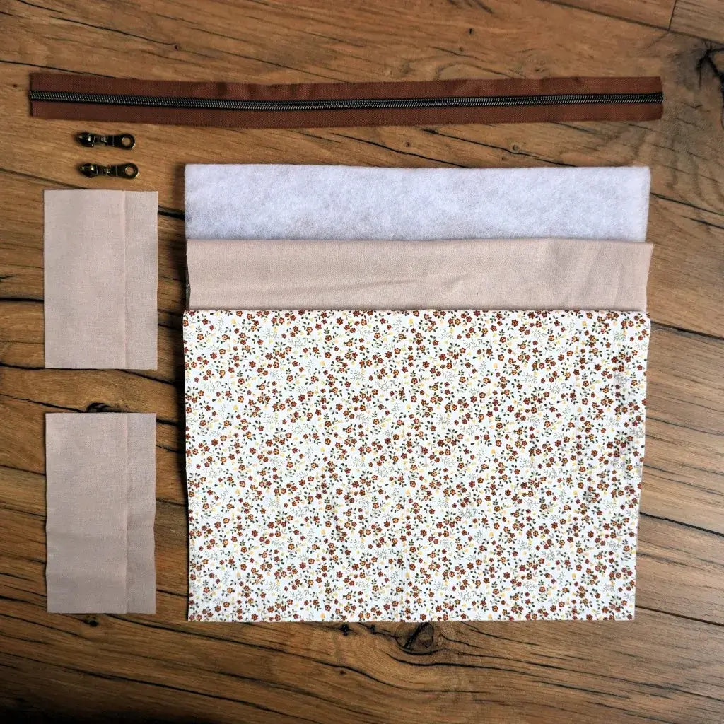 Kit couture complet pour réaliser une trousse carrée à motif fleuri façon prairie, composé du tissu principal à petites fleurs, d’une doublure beige, de pièces de ouate, d’une fermeture éclair marron et de deux curseurs métalliques. Ce kit prêt-à-coudre permet de fabriquer facilement une trousse personnalisée au style champêtre, idéale pour offrir ou organiser ses accessoires de toilette.