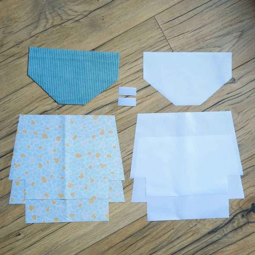 Kit de couture complet pour fabriquer une pochette à scratch imperméable, composé d’un rabat en velours côtelé bleu, de plusieurs pièces de coton imprimé aux motifs géométriques façon terrazzo bleu et ocre, de doublures blanches en coton enduit et de deux bandes de scratch. Les éléments sont soigneusement disposés au sol, prêts à être assemblés pour créer une trousse pratique et personnalisée.