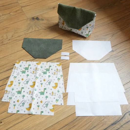 Kit couture pour réaliser une pochette à scratch imperméable, comprenant plusieurs pièces de tissu prédécoupées : coton imprimé dinosaures colorés, velours côtelé vert pour le rabat, doublures blanches en coton enduit et scratchs blancs. Les éléments sont disposés au sol à côté d’un modèle déjà cousu, idéal pour apprendre à assembler facilement une trousse pratique et personnalisée.