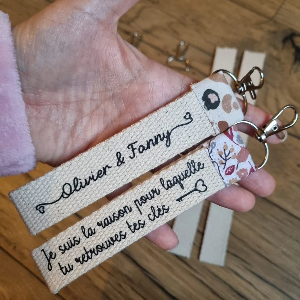 Duo de porte-clés avec inscription personnalisée présentés dans la paume d'une main. Le premier porte-clé est personnalisé avec l'inscription "Olivier & Fanny", le second avec l'inscription "Je suis la raison pour laquelle tu retrouves tes clés". A l'arrière-plan, on distingue les différentes pièces nécessaires à la confection de ce duo de porte-clés personnalisés.