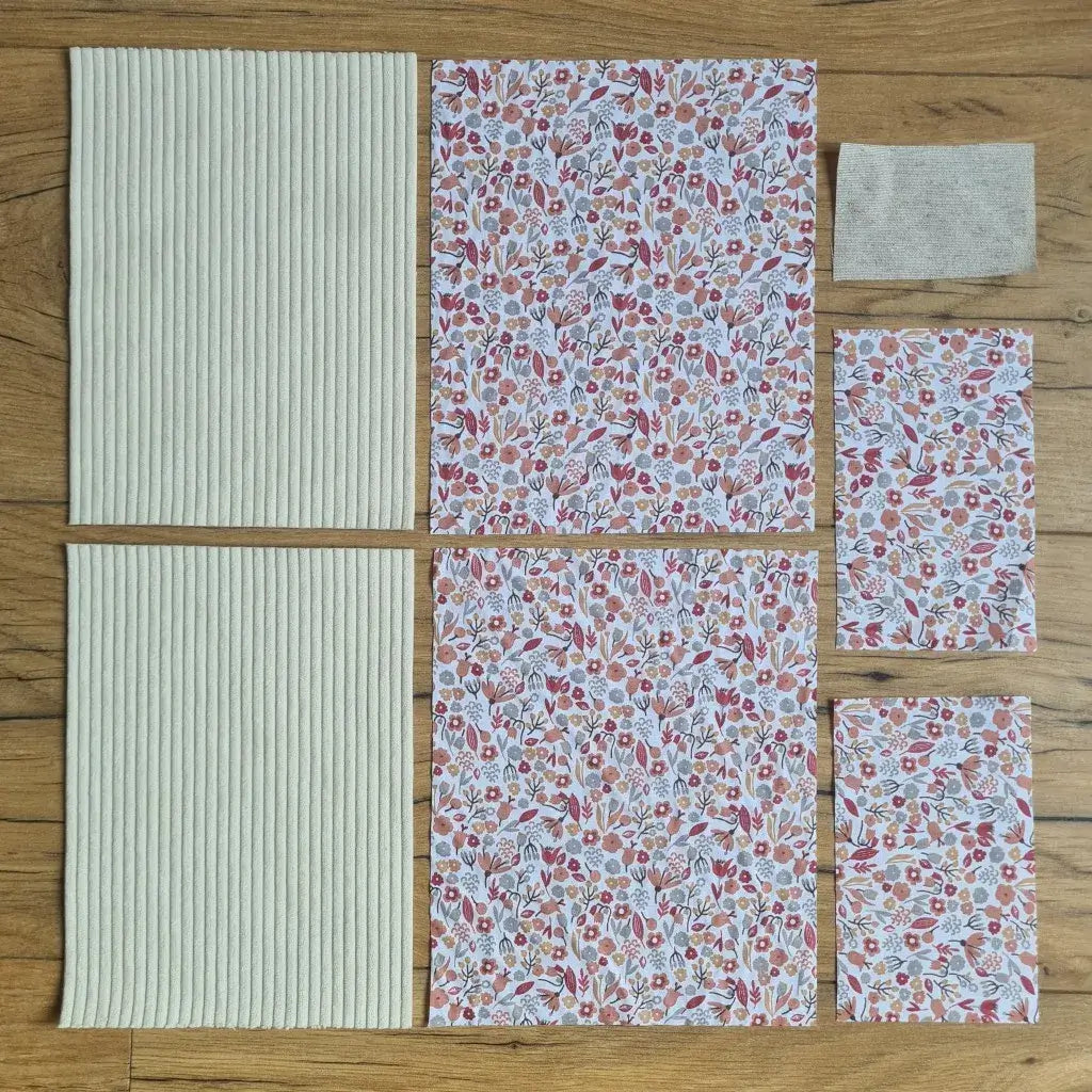 Kit "prêt à coudre" pour une pochette à livre & liseuse en velours côtelé au coloris beige avec une doublure en tissu à motif liberty. L'ensemble des pièces de tissu, déjà prédécoupées aux bonnes dimensions, sont disposées au sol. A gauche, deux pièces identiques en velours côtelé beige. A droite, quatre pièces en tissu imprimé à motif liberty ainsi qu’une étiquette en tissu toile uni servant de support à une inscription personnalisée qui sera floquée dessus.