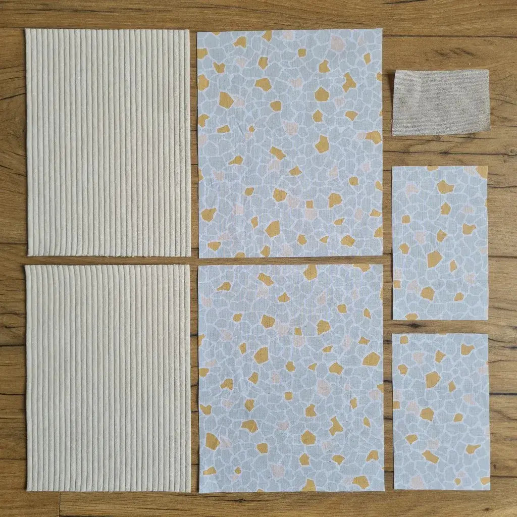 Kit "prêt à coudre" pour une pochette à livre & liseuse en velours côtelé au coloris beige avec une doublure en tissu à motif argeo. L'ensemble des pièces de tissu, déjà prédécoupées aux bonnes dimensions, sont disposées au sol. A gauche, deux pièces identiques en velours côtelé beige. A droite, quatre pièces en tissu imprimé à motif argeo ainsi qu’une étiquette en tissu toile uni servant de support à une inscription personnalisée qui sera floquée dessus.