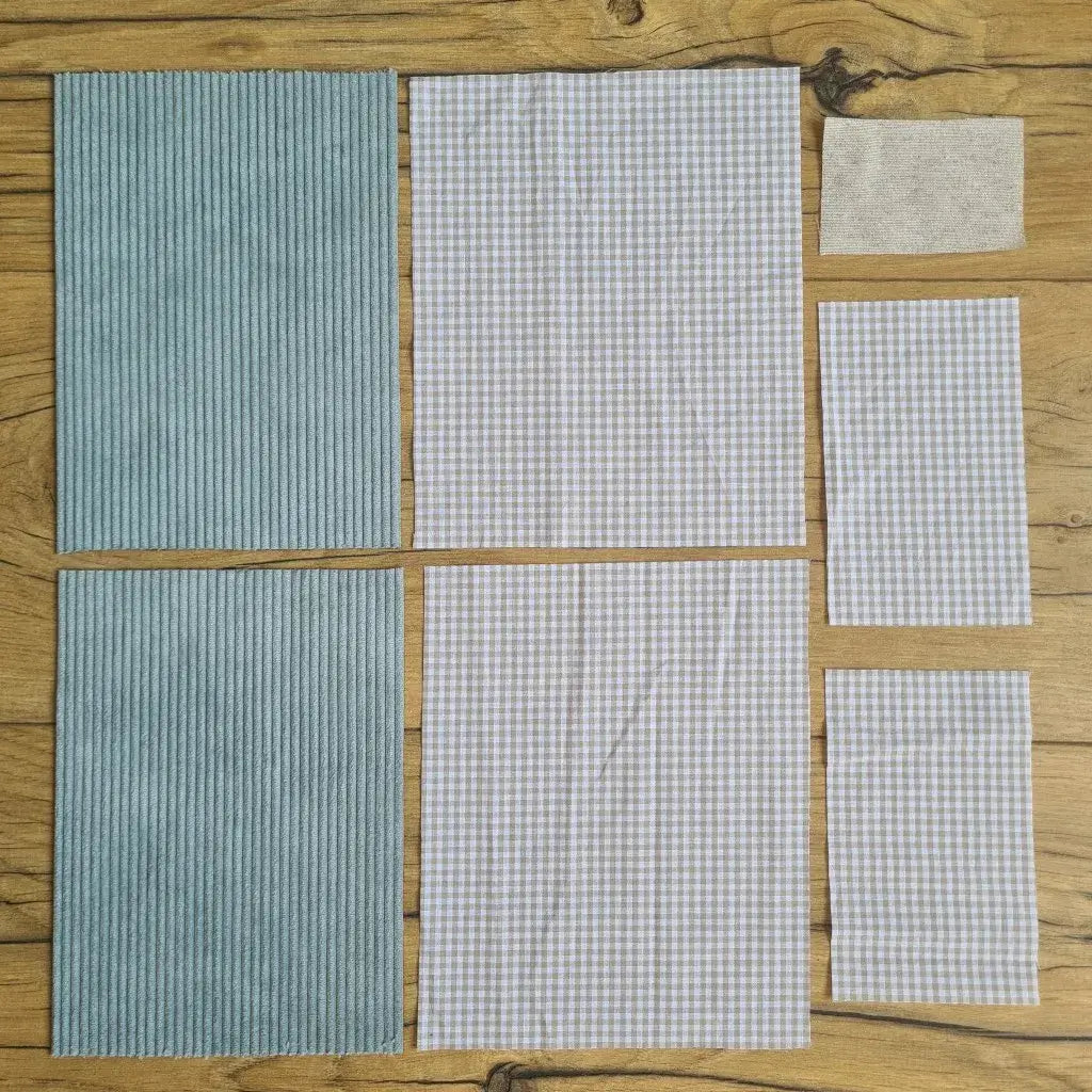 Kit "prêt à coudre" pour une pochette à livre & liseuse en velours côtelé au coloris bleu clair avec une doublure en tissu à motif carreaux beiges. L'ensemble des pièces de tissu, déjà prédécoupées aux bonnes dimensions, sont disposées au sol. A gauche, deux pièces identiques en velours côtelé bleu clair. A droite, quatre pièces en tissu imprimé à motif carreaux beiges ainsi qu’une étiquette en tissu toile uni servant de support à une inscription personnalisée qui sera floquée dessus.