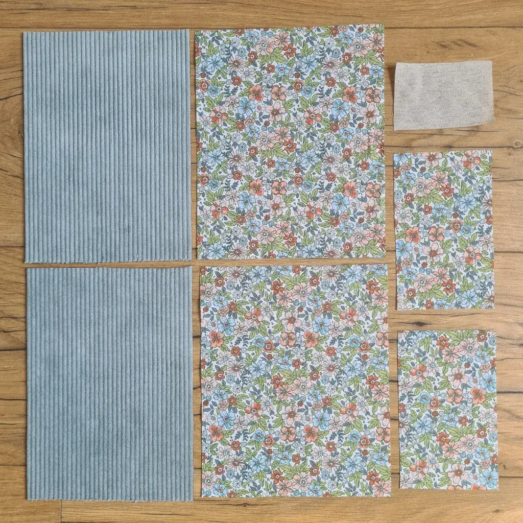 Kit "prêt à coudre" pour une pochette à livre & liseuse en velours côtelé au coloris bleu clair avec une doublure en tissu à motif fleuri. L'ensemble des pièces de tissu, déjà prédécoupées aux bonnes dimensions, sont disposées au sol. A gauche, deux pièces identiques en velours côtelé bleu clair. A droite, quatre pièces en tissu imprimé à motif fleuri ainsi qu’une étiquette en tissu toile uni servant de support à une inscription personnalisée qui sera floquée dessus.