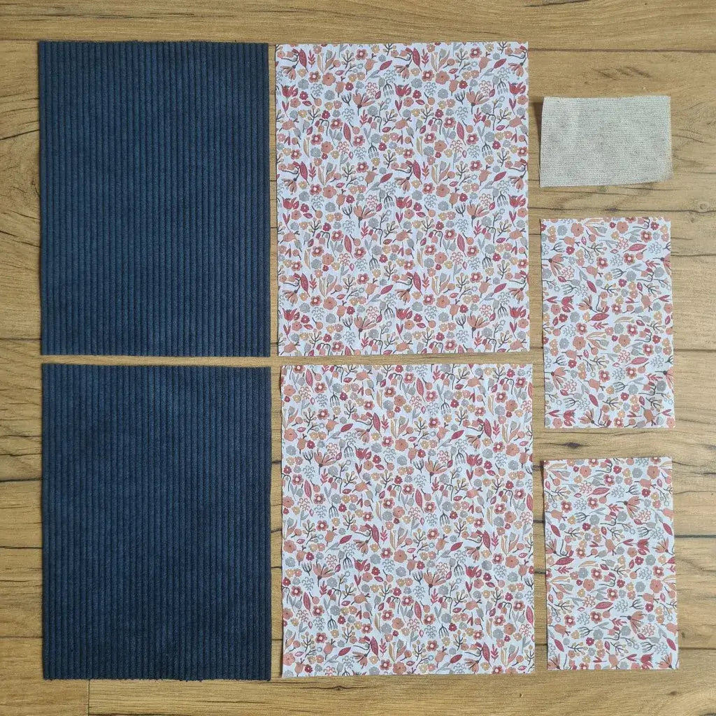 Kit "prêt à coudre" pour une pochette à livre & liseuse en velours côtelé au coloris bleu foncé avec une doublure en tissu à motif liberty. L'ensemble des pièces de tissu, déjà prédécoupées aux bonnes dimensions, sont disposées au sol. A gauche, deux pièces identiques en velours côtelé bleu foncé. A droite, quatre pièces en tissu imprimé à motif liberty ainsi qu’une étiquette en tissu toile uni servant de support à une inscription personnalisée qui sera floquée dessus.