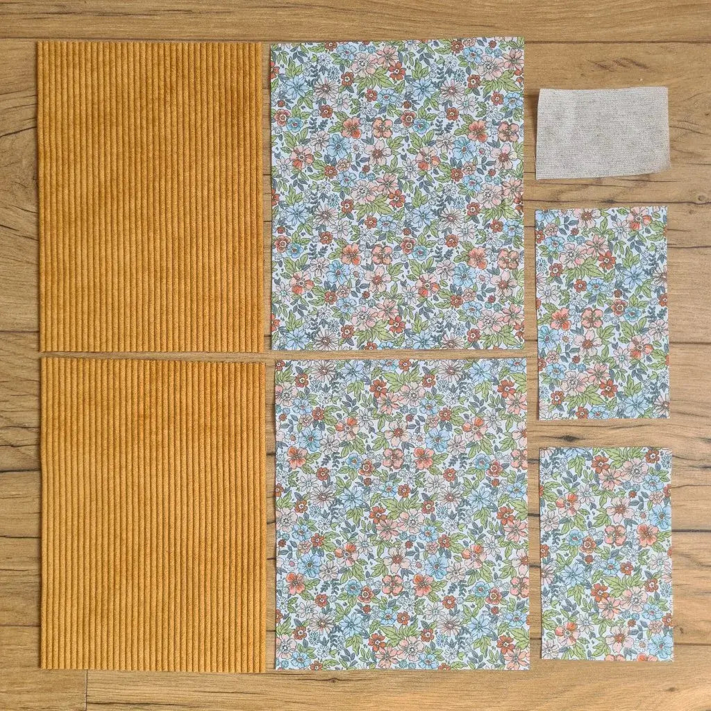 Kit "prêt à coudre" pour une pochette à livre & liseuse en velours côtelé au coloris jaune avec une doublure en tissu à motif fleuri. L'ensemble des pièces de tissu, déjà prédécoupées aux bonnes dimensions, sont disposées au sol. A gauche, deux pièces identiques en velours côtelé jaune. A droite, quatre pièces en tissu imprimé à motif fleuri ainsi qu’une étiquette en tissu toile uni servant de support à une inscription personnalisée qui sera floquée dessus.