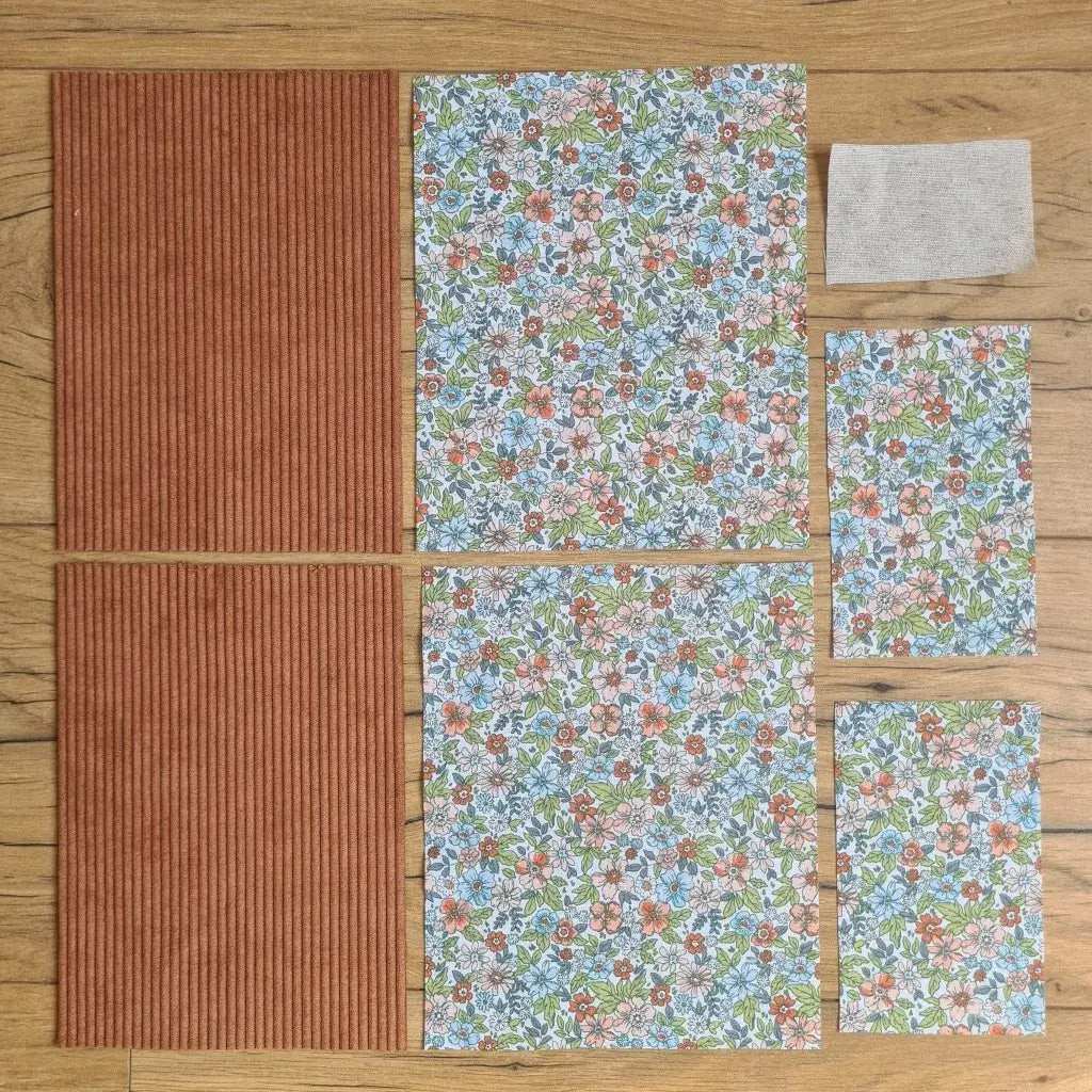 Kit "prêt à coudre" pour une pochette à livre & liseuse en velours côtelé au coloris marron avec une doublure en tissu à motif fleuri. L'ensemble des pièces de tissu, déjà prédécoupées aux bonnes dimensions, sont disposées au sol. A gauche, deux pièces identiques en velours côtelé marron. A droite, quatre pièces en tissu imprimé à motif fleuri ainsi qu’une étiquette en tissu toile uni servant de support à une inscription personnalisée qui sera floquée dessus.