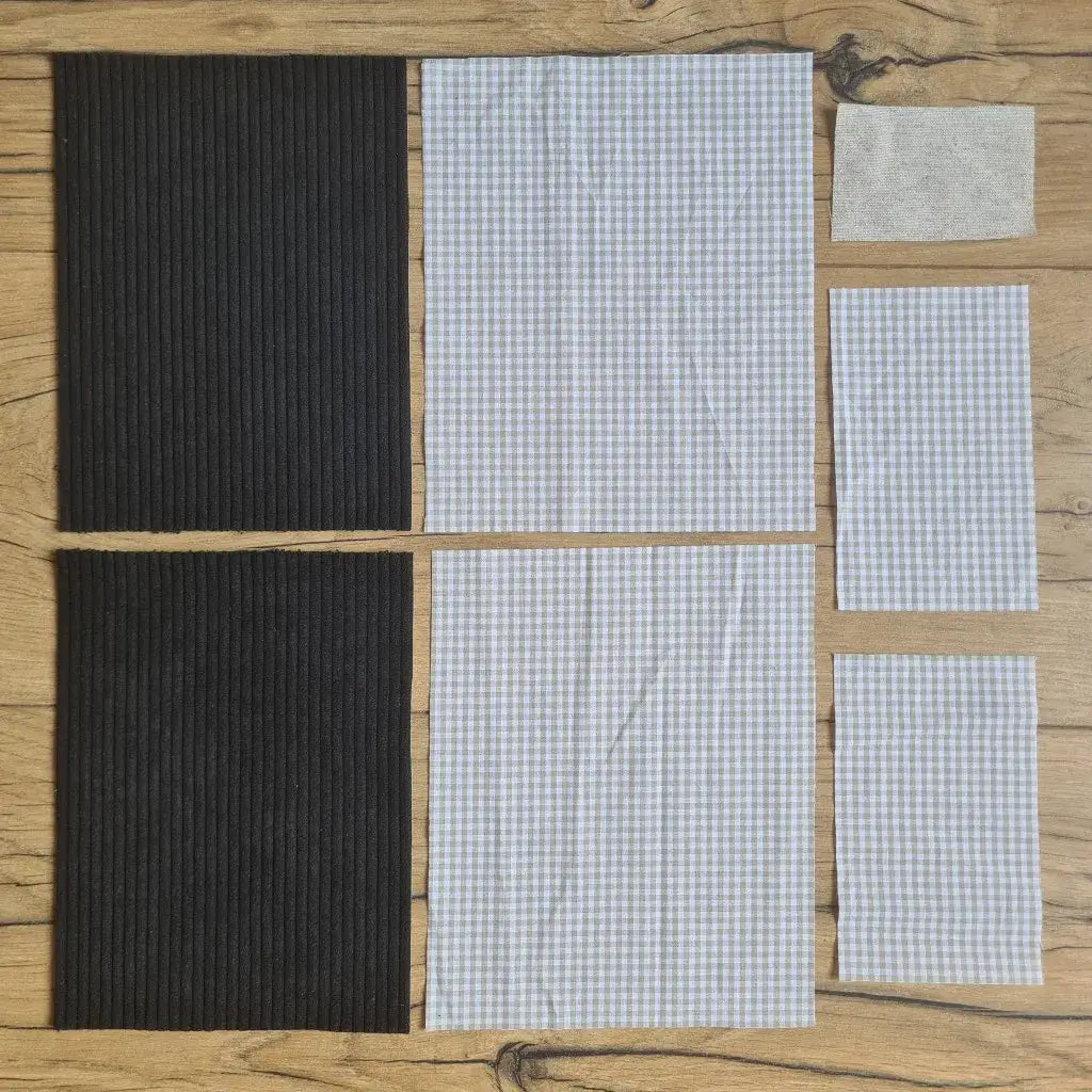 Kit "prêt à coudre" pour une pochette à livre & liseuse en velours côtelé au coloris noir avec une doublure en tissu à motif carreaux beiges. L'ensemble des pièces de tissu, déjà prédécoupées aux bonnes dimensions, sont disposées au sol. A gauche, deux pièces identiques en velours côtelé noir. A droite, quatre pièces en tissu imprimé à motif carreaux beiges ainsi qu’une étiquette en tissu toile uni servant de support à une inscription personnalisée qui sera floquée dessus.