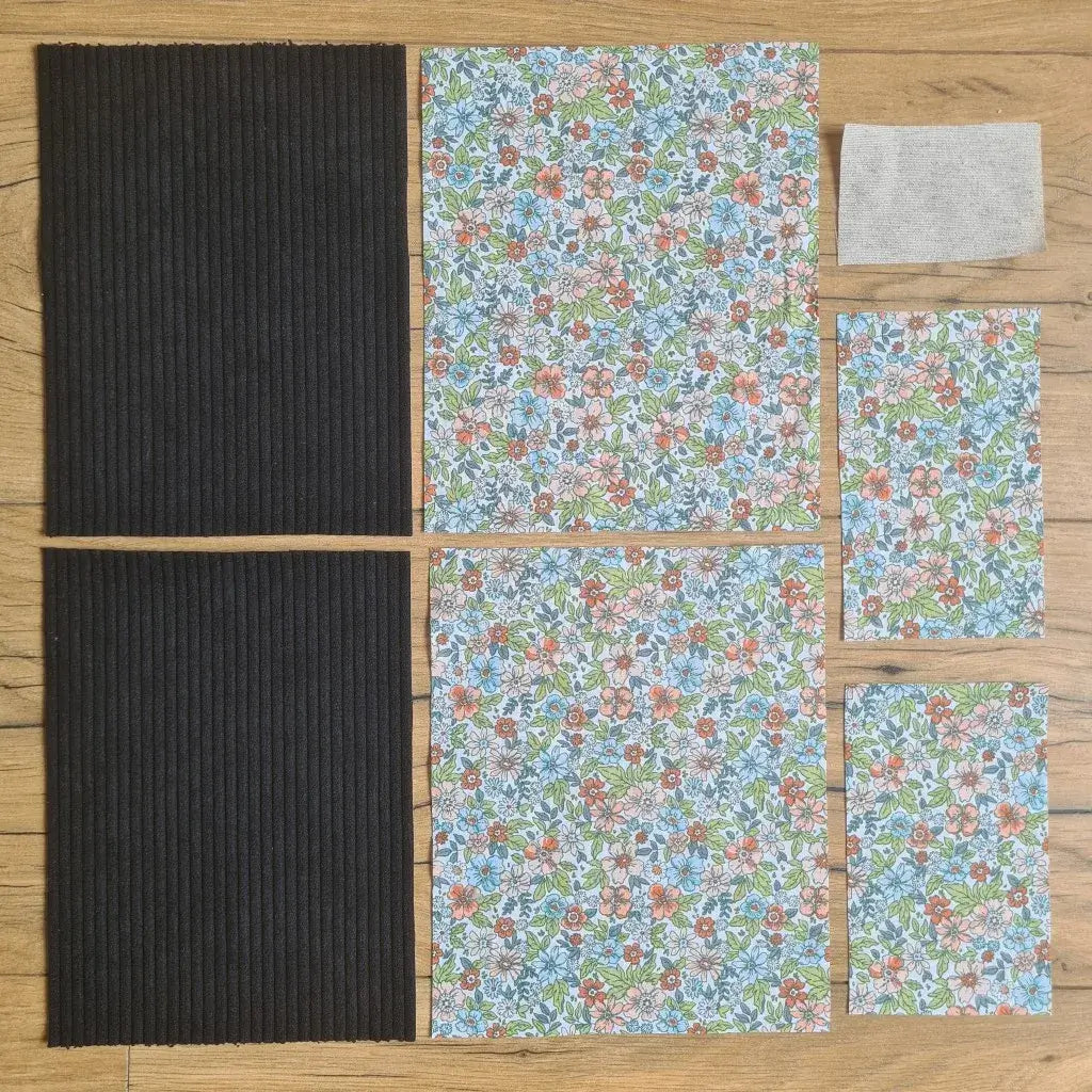 Kit "prêt à coudre" pour une pochette à livre & liseuse en velours côtelé au coloris noir avec une doublure en tissu à motif fleuri. L'ensemble des pièces de tissu, déjà prédécoupées aux bonnes dimensions, sont disposées au sol. A gauche, deux pièces identiques en velours côtelé noir. A droite, quatre pièces en tissu imprimé à motif fleuri ainsi qu’une étiquette en tissu toile uni servant de support à une inscription personnalisée qui sera floquée dessus.