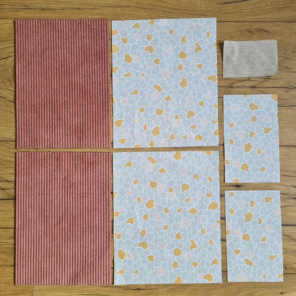 Kit "prêt à coudre" pour une pochette à livre & liseuse en velours côtelé au coloris rose avec une doublure en tissu à motif argeo. L'ensemble des pièces de tissu, déjà prédécoupées aux bonnes dimensions, sont disposées au sol. A gauche, deux pièces identiques en velours côtelé rose. A droite, quatre pièces en tissu imprimé à motif argeo ainsi qu’une étiquette en tissu toile uni servant de support à une inscription personnalisée qui sera floquée dessus.