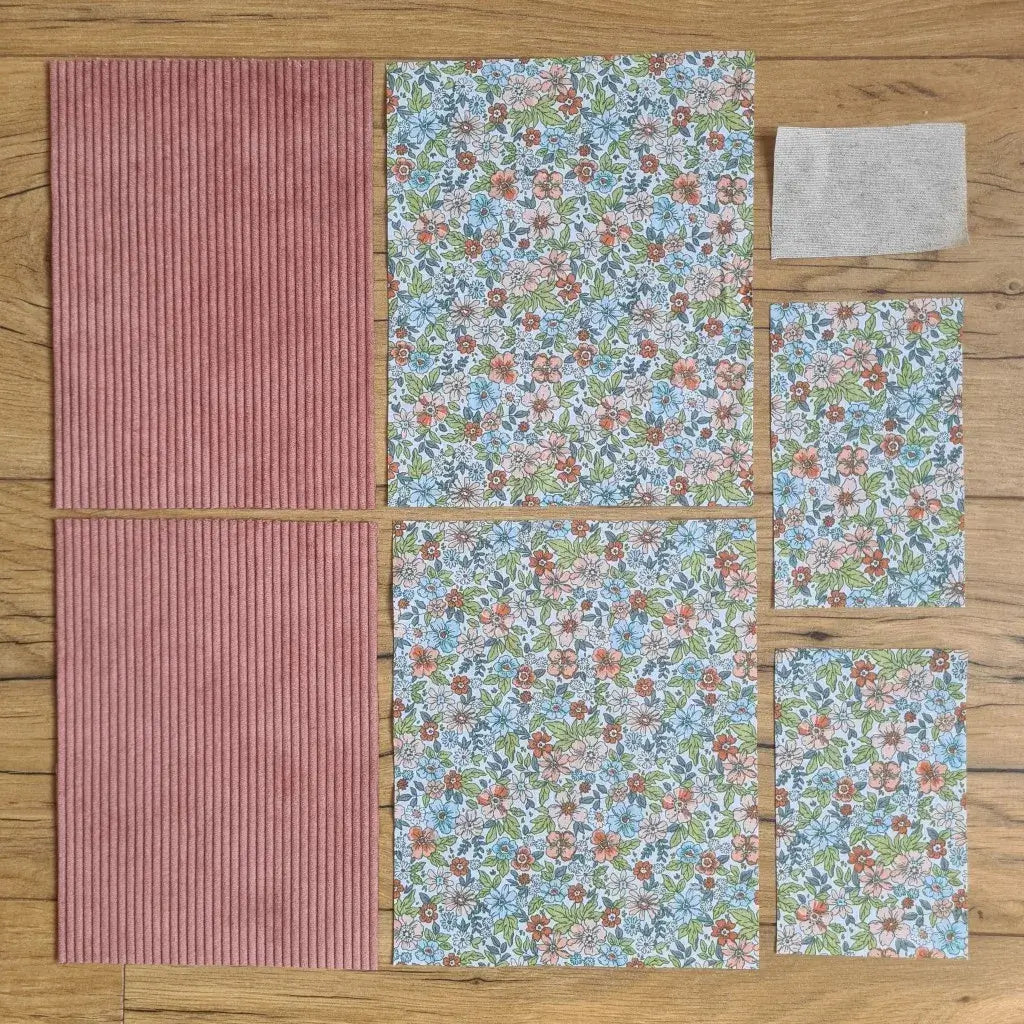 Kit "prêt à coudre" pour une pochette à livre & liseuse en velours côtelé au coloris rose avec une doublure en tissu à motif fleuri. L'ensemble des pièces de tissu, déjà prédécoupées aux bonnes dimensions, sont disposées au sol. A gauche, deux pièces identiques en velours côtelé rose. A droite, quatre pièces en tissu imprimé à motif fleuri ainsi qu’une étiquette en tissu toile uni servant de support à une inscription personnalisée qui sera floquée dessus.