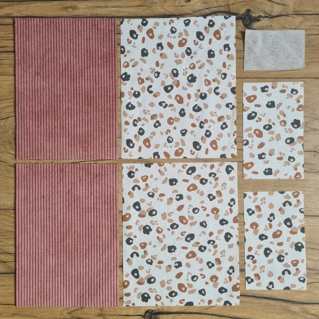 Kit "prêt à coudre" pour une pochette à livre & liseuse en velours côtelé au coloris rose avec une doublure en tissu à motif léopard. L'ensemble des pièces de tissu, déjà prédécoupées aux bonnes dimensions, sont disposées au sol. A gauche, deux pièces identiques en velours côtelé rose. A droite, quatre pièces en tissu imprimé à motif léopard ainsi qu’une étiquette en tissu toile uni servant de support à une inscription personnalisée qui sera floquée dessus.