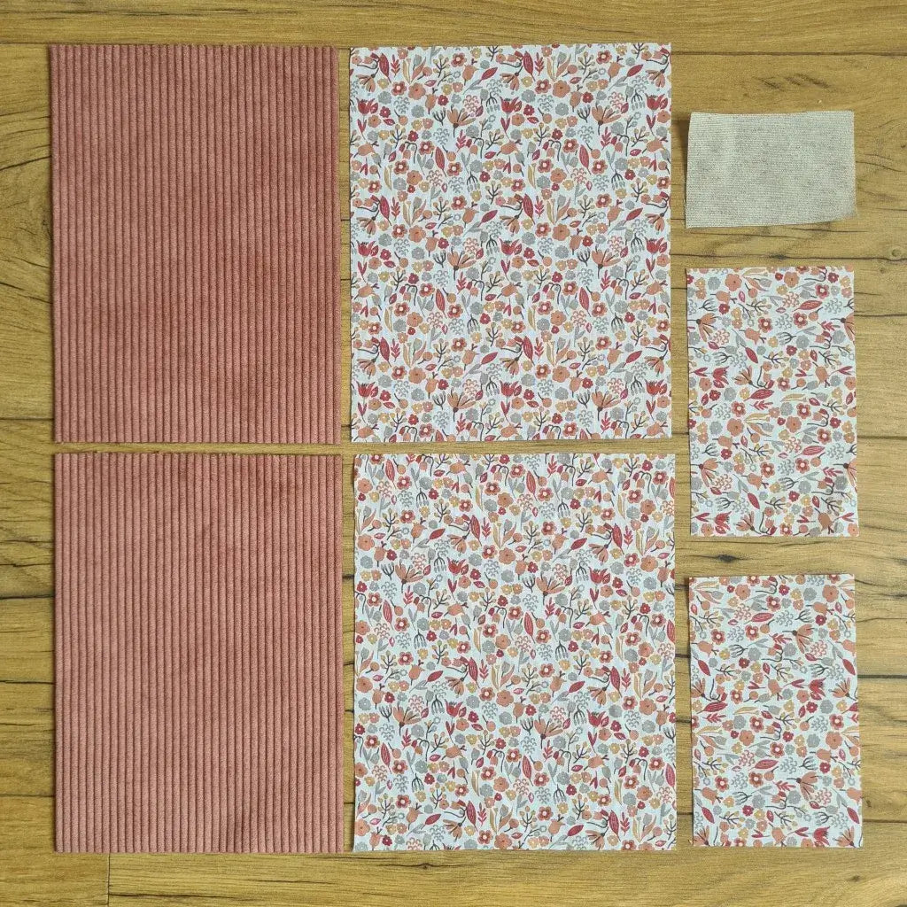 Kit "prêt à coudre" pour une pochette à livre & liseuse en velours côtelé au coloris rose avec une doublure en tissu à motif liberty. L'ensemble des pièces de tissu, déjà prédécoupées aux bonnes dimensions, sont disposées au sol. A gauche, deux pièces identiques en velours côtelé rose. A droite, quatre pièces en tissu imprimé à motif liberty ainsi qu’une étiquette en tissu toile uni servant de support à une inscription personnalisée qui sera floquée dessus.