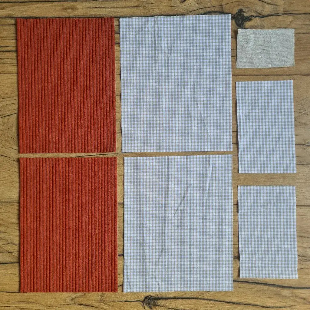 Kit "prêt à coudre" pour une pochette à livre & liseuse en velours côtelé au coloris terracotta avec une doublure en tissu à motif carreaux beiges. L'ensemble des pièces de tissu, déjà prédécoupées aux bonnes dimensions, sont disposées au sol. A gauche, deux pièces identiques en velours côtelé terracotta. A droite, quatre pièces en tissu imprimé à motif carreaux beiges ainsi qu’une étiquette en tissu toile uni servant de support à une inscription personnalisée qui sera floquée dessus.