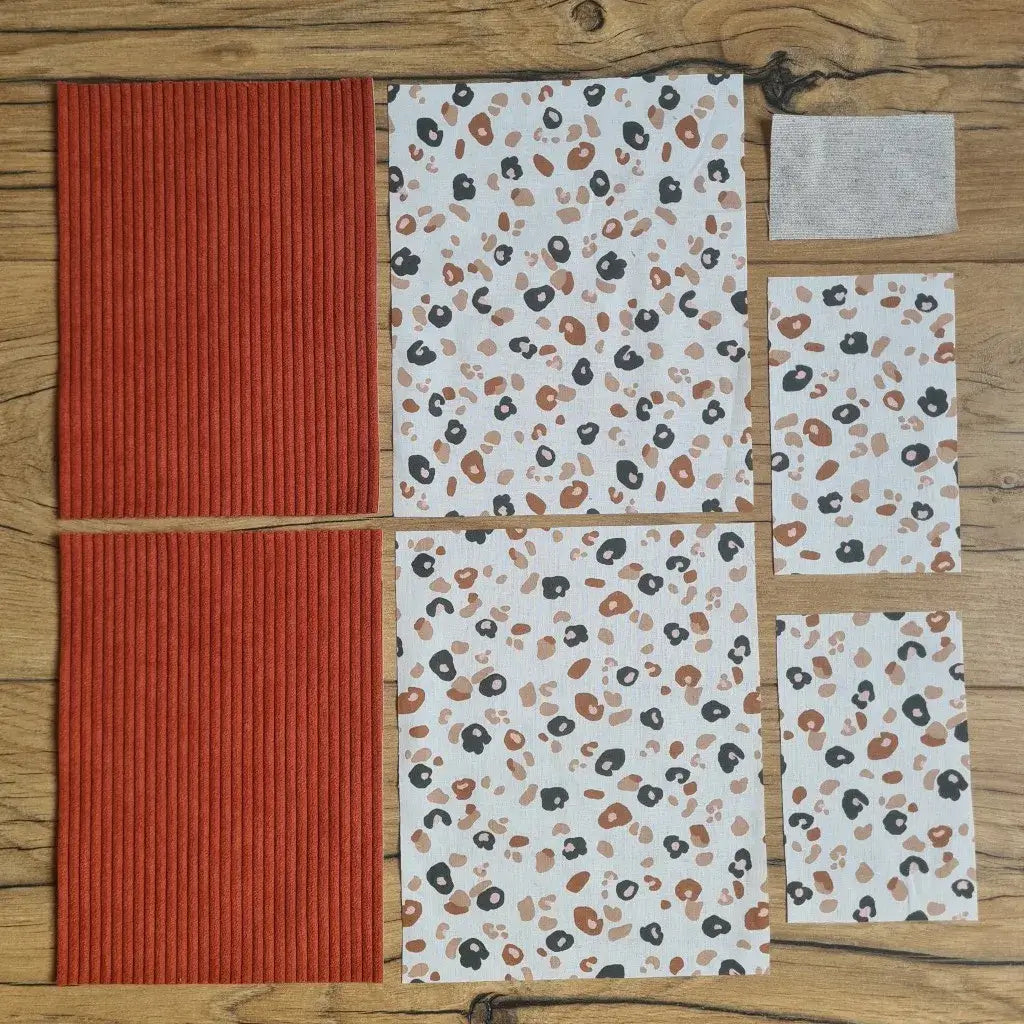 Kit "prêt à coudre" pour une pochette à livre & liseuse en velours côtelé au coloris terracotta avec une doublure en tissu à motif léopard. L'ensemble des pièces de tissu, déjà prédécoupées aux bonnes dimensions, sont disposées au sol. A gauche, deux pièces identiques en velours côtelé terracotta. A droite, quatre pièces en tissu imprimé à motif léopard ainsi qu’une étiquette en tissu toile uni servant de support à une inscription personnalisée qui sera floquée dessus.