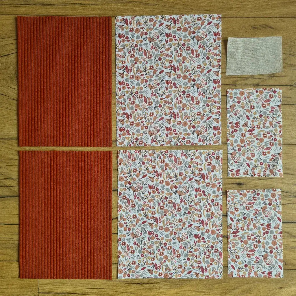 Kit "prêt à coudre" pour une pochette à livre & liseuse en velours côtelé au coloris terracotta avec une doublure en tissu à motif liberty. L'ensemble des pièces de tissu, déjà prédécoupées aux bonnes dimensions, sont disposées au sol. A gauche, deux pièces identiques en velours côtelé terracotta. A droite, quatre pièces en tissu imprimé à motif liberty ainsi qu’une étiquette en tissu toile uni servant de support à une inscription personnalisée qui sera floquée dessus.