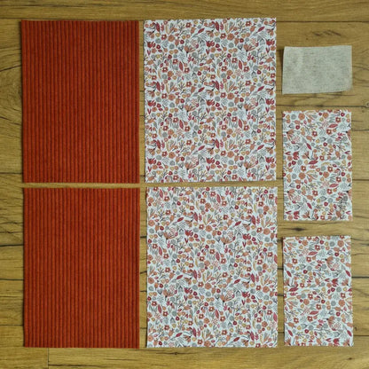 Kit "prêt à coudre" pour une pochette à livre & liseuse en velours côtelé au coloris terracotta avec une doublure en tissu à motif liberty. L'ensemble des pièces de tissu, déjà prédécoupées aux bonnes dimensions, sont disposées au sol. A gauche, deux pièces identiques en velours côtelé terracotta. A droite, quatre pièces en tissu imprimé à motif liberty ainsi qu’une étiquette en tissu toile uni servant de support à une inscription personnalisée qui sera floquée dessus.