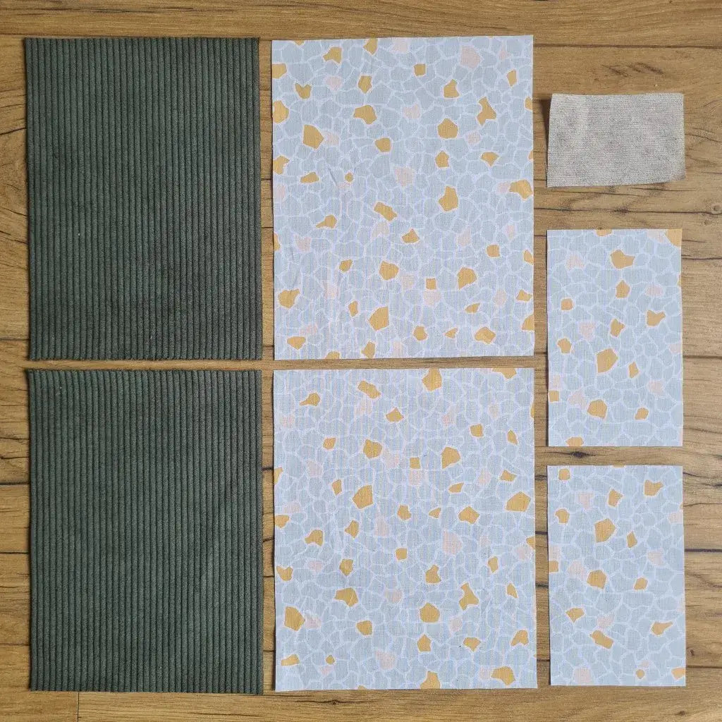 Kit "prêt à coudre" pour une pochette à livre & liseuse en velours côtelé au coloris vert avec une doublure en tissu à motif argeo. L'ensemble des pièces de tissu, déjà prédécoupées aux bonnes dimensions, sont disposées au sol. A gauche, deux pièces identiques en velours côtelé vert. A droite, quatre pièces en tissu imprimé à motif argeo ainsi qu’une étiquette en tissu toile uni servant de support à une inscription personnalisée qui sera floquée dessus.