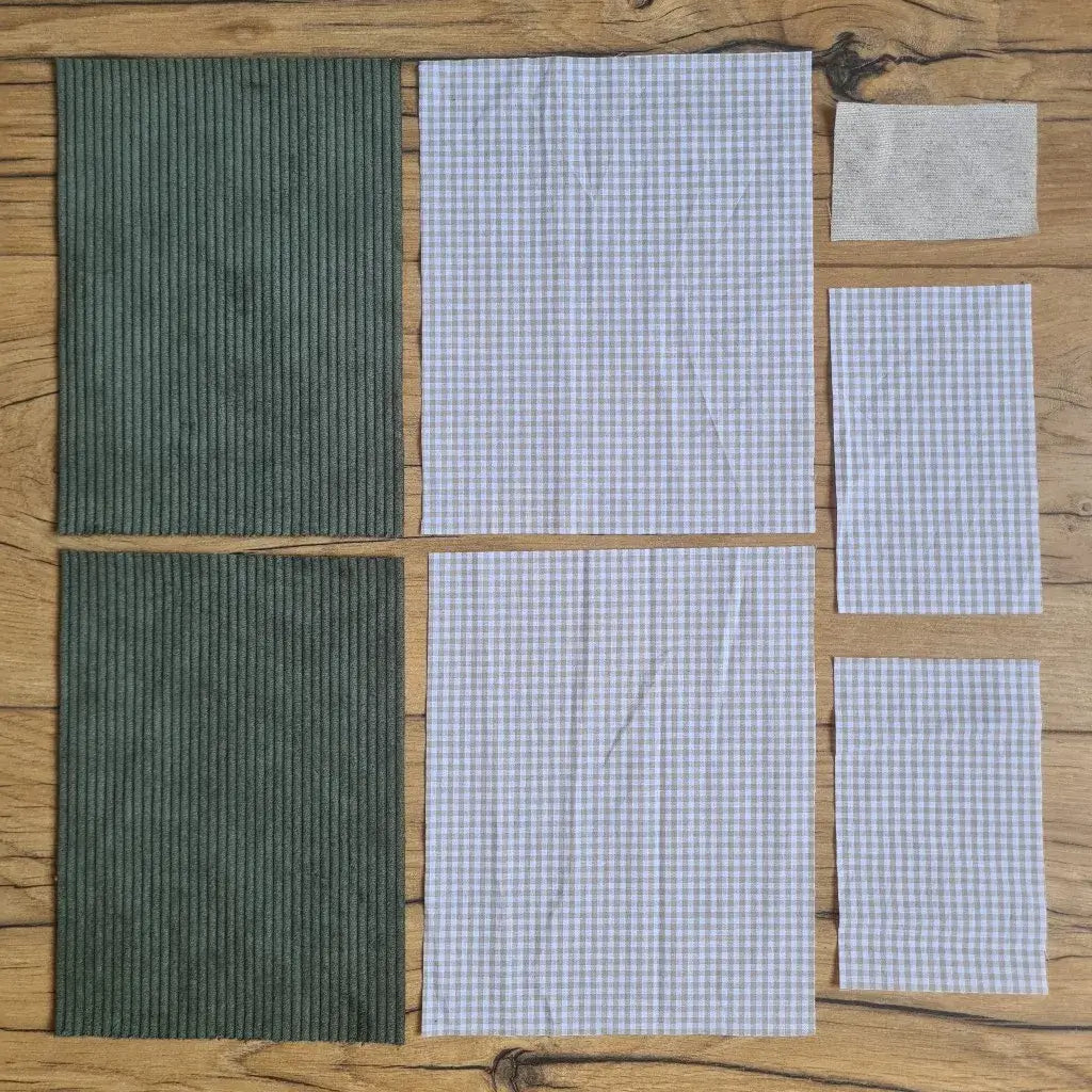 Kit "prêt à coudre" pour une pochette à livre & liseuse en velours côtelé au coloris vert avec une doublure en tissu à motif carreaux beiges. L'ensemble des pièces de tissu, déjà prédécoupées aux bonnes dimensions, sont disposées au sol. A gauche, deux pièces identiques en velours côtelé vert. A droite, quatre pièces en tissu imprimé à motif carreaux beiges ainsi qu’une étiquette en tissu toile uni servant de support à une inscription personnalisée qui sera floquée dessus.