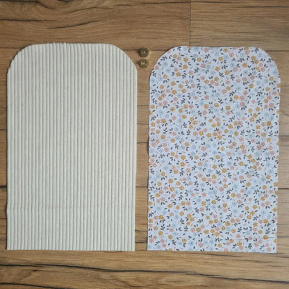 Kit "prêt à coudre" pour une pochette aimantée en velours côtelé au coloris beige avec une doublure en tissu à motif fleurette. L'ensemble des pièces de tissu, déjà prédécoupées aux bonnes dimensions, sont disposées au sol. A gauche, une pièce en velours côtelé beige. A droite, une pièce en tissu imprimé à motif fleurette. Entre les deux, les 2 pièces composant un bouton aimanté.