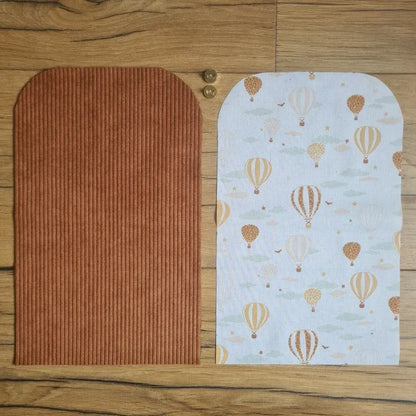 Kit "prêt à coudre" pour une pochette aimantée en velours côtelé au coloris marron avec une doublure en tissu à motif montgolfières. L'ensemble des pièces de tissu, déjà prédécoupées aux bonnes dimensions, sont disposées au sol. A gauche, une pièce en velours côtelé marron. A droite, une pièce en tissu imprimé à motif montgolfières. Entre les deux, les 2 pièces composant un bouton aimanté.