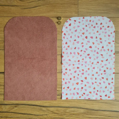 Kit "prêt à coudre" pour une pochette aimantée en velours côtelé au coloris rose avec une doublure en tissu à motif cœurs. L'ensemble des pièces de tissu, déjà prédécoupées aux bonnes dimensions, sont disposées au sol. A gauche, une pièce en velours côtelé rose. A droite, une pièce en tissu imprimé à motif cœurs. Entre les deux, les 2 pièces composant un bouton aimanté.