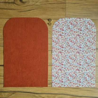 Kit "prêt à coudre" pour une pochette aimantée en velours côtelé au coloris terracotta avec une doublure en tissu à motif liberty. L'ensemble des pièces de tissu, déjà prédécoupées aux bonnes dimensions, sont disposées au sol. A gauche, une pièce en velours côtelé terracotta. A droite, une pièce en tissu imprimé à motif liberty. Entre les deux, les 2 pièces composant un bouton aimanté.