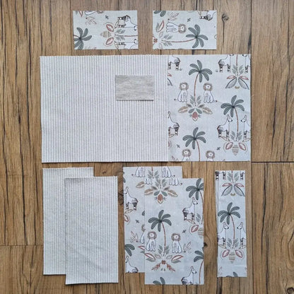 Kit prêt-à-coudre pour protège carnet de santé, composé de tissus prédécoupés avec un motif jungle et un velours côtelé beige, présenté à plat sur un sol en bois. Accessoire DIY idéal pour confectionner un protège carnet unique et personnalisé. Motif exotique avec lions, girafes et palmiers, parfait pour un cadeau de naissance ou pour organiser le carnet de bébé avec style. Kit comprenant toutes les pièces nécessaires pour une création artisanale facile et soignée.