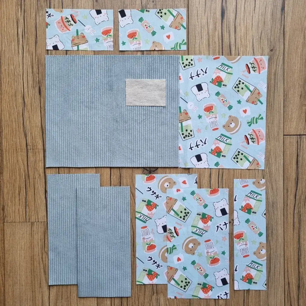 Kit prêt-à-coudre pour protège carnet de santé, comprenant des tissus prédécoupés avec un motif kawaii et un velours côtelé bleu clair, présenté à plat sur un sol en bois. Accessoire DIY idéal pour confectionner un protège carnet unique et personnalisé. Imprimé ludique avec animaux, sushi, bubble tea et fraises, parfait pour un cadeau de naissance original ou pour organiser le carnet de bébé avec une touche fun. Kit complet avec toutes les pièces nécessaires pour une création artisanale facile et soignée.