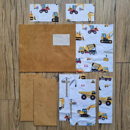 Kit prêt-à-coudre pour protège carnet de santé, composé de tissus prédécoupés en velours côtelé jaune moutarde et d’un imprimé chantier avec camions, grues et pelleteuses, présenté à plat sur un sol en bois. Accessoire DIY idéal pour confectionner un protège carnet unique et personnalisé. Modèle avec rabats et lien de fermeture, parfait pour organiser le carnet de bébé ou offrir un cadeau de naissance original. Kit complet avec toutes les pièces nécessaires pour une couture facile et un rendu soigné.