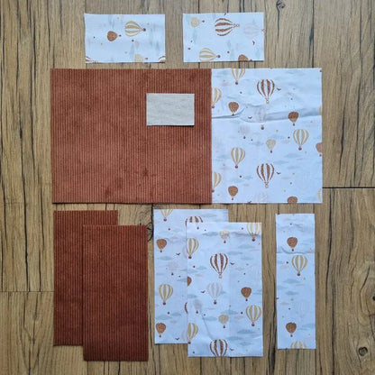 Kit prêt-à-coudre pour protège carnet de santé, comprenant des tissus prédécoupés en velours côtelé marron et un imprimé montgolfières, présenté à plat sur un sol en bois. Accessoire DIY idéal pour confectionner un protège carnet unique et personnalisé. Motif poétique avec montgolfières et nuages, parfait pour un cadeau de naissance ou pour organiser le carnet de bébé avec élégance. Kit complet avec toutes les pièces nécessaires pour une couture facile et un rendu soigné.
