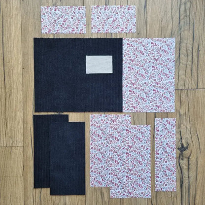 Kit prêt-à-coudre pour protège carnet de santé, comprenant des tissus prédécoupés en velours côtelé noir et un imprimé floral liberty rose et blanc, présenté à plat sur un sol en bois. Accessoire DIY idéal pour créer un protège carnet unique et élégant. Modèle avec rabats et lien de fermeture, parfait pour organiser le carnet de bébé ou offrir un cadeau de naissance personnalisé. Kit complet avec toutes les pièces nécessaires pour une couture facile et un rendu soigné.