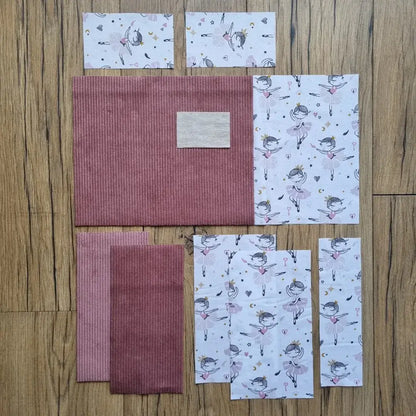 Kit prêt-à-coudre pour protège carnet de santé, composé de tissus prédécoupés en velours côtelé rose poudré et d’un imprimé danseuses étoile sur fond blanc, présenté à plat sur un sol en bois. Accessoire DIY idéal pour confectionner un protège carnet unique et élégant. Motif féérique avec ballerines, cœurs et étoiles, parfait pour un cadeau de naissance ou pour organiser le carnet de bébé avec une touche poétique. Kit complet avec toutes les pièces nécessaires pour une couture facile et un rendu soigné.
