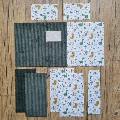 Kit prêt-à-coudre pour protège carnet de santé, comprenant des tissus prédécoupés en velours côtelé vert et un imprimé dinosaures sur fond blanc, présenté à plat sur un sol en bois. Accessoire DIY idéal pour confectionner un protège carnet unique et original. Motif ludique avec dinosaures et feuillages, parfait pour un cadeau de naissance ou pour organiser le carnet de bébé avec une touche amusante. Kit complet avec toutes les pièces nécessaires pour une couture facile et un rendu soigné.