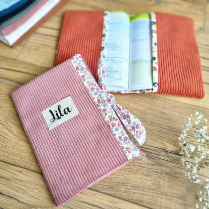 Kit prêt-à-coudre pour protège carnet de santé en velours côtelé avec intérieur fleuri, présenté sur un sol en bois. Accessoire personnalisable, idéal pour protéger et embellir un carnet de santé tout en ajoutant une touche unique. Modèle avec rabats et lien de fermeture, disponible en plusieurs coloris, dont rose poudré et terracotta. Fabrication artisanale avec finitions soignées, parfait pour un cadeau de naissance ou une organisation pratique du carnet de bébé.